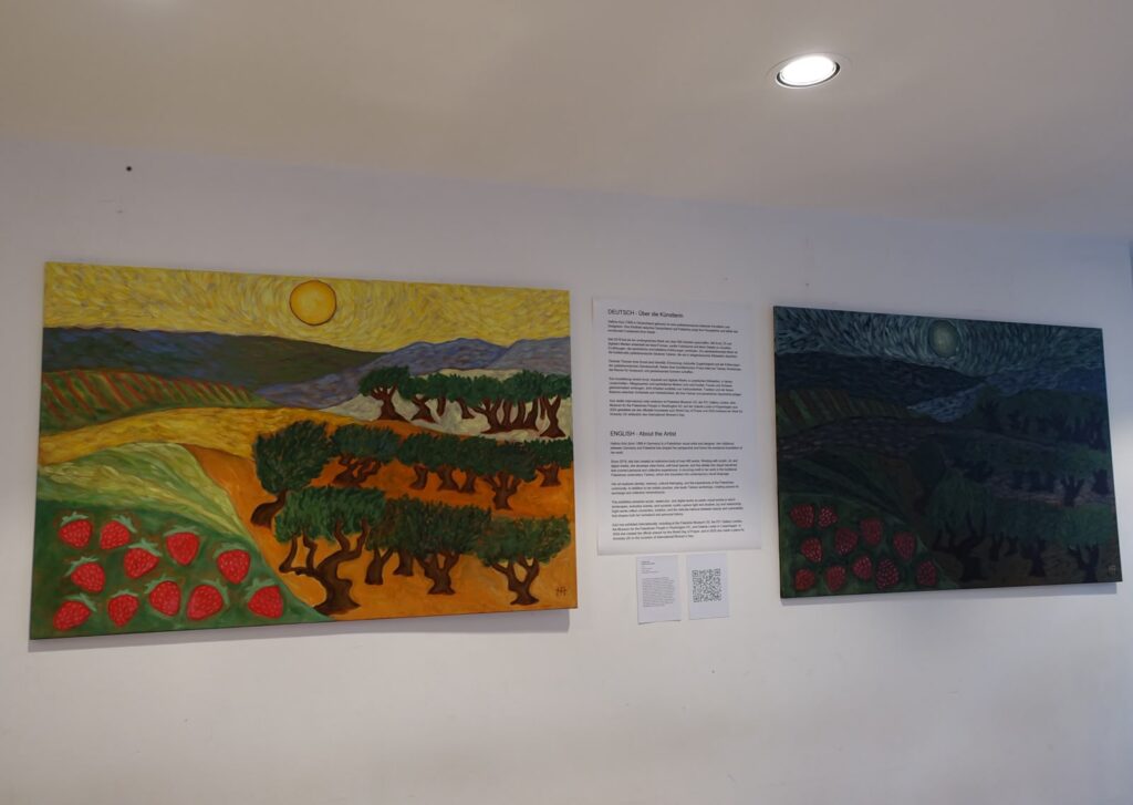 Zwei Landschaftsgemälde mit Olivenbäumen und Erdbeeren hängen in einer Ausstellung an einer weißen Wand, daneben ein Infotext